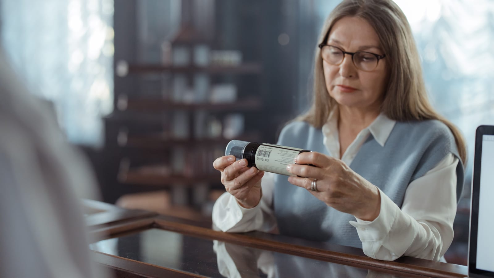 Older woman in glasses reading a prescription bottle label at a pharmacy counter